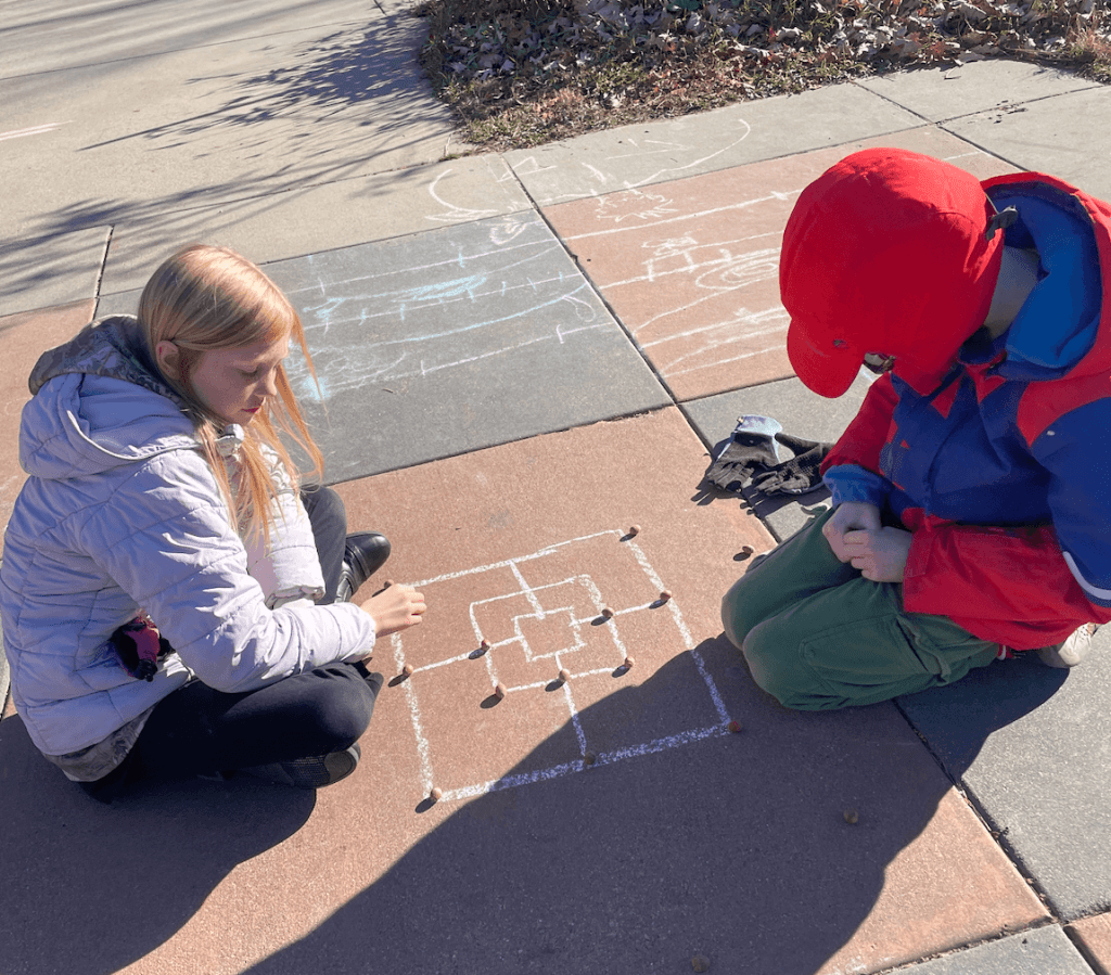two children playing a game with chalk and pebbles outside